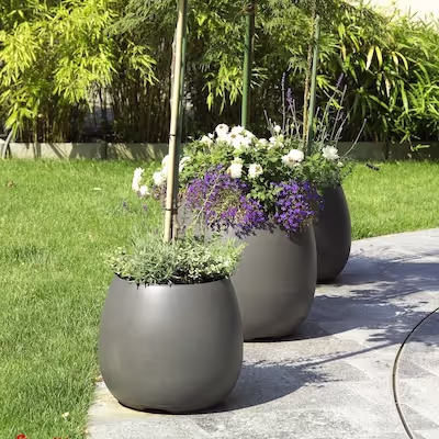 Drei Swisspearl Sumo Pflanzgefäße mit Bäumen und Blumen im Garten.